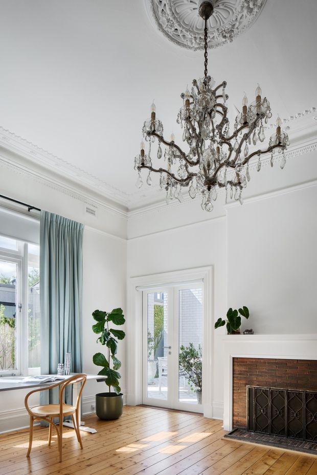 A bright, elegant heritage-style living room or dining area with white walls, ornate ceiling cornices and a decorative ceiling medallion, a large crystal chandelier, timber floorboards, tall windows with pale blue curtains, and a traditional brick fireplace with a decorative metal screen. A small table and bentwood chairs occupy the window side, while potted plants add greenery.