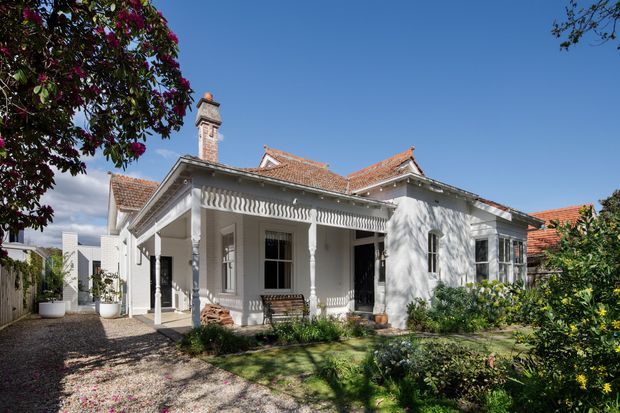 A single-storey heritage-style residence viewed from the front garden, featuring a white rendered facade, ornate Victorian-era verandah with decorative turned posts and fretwork, a prominent brick chimney, and multiple terracotta tiled gabled roof sections. The home is set within established landscaped gardens and a gravel forecourt under clear blue skies.