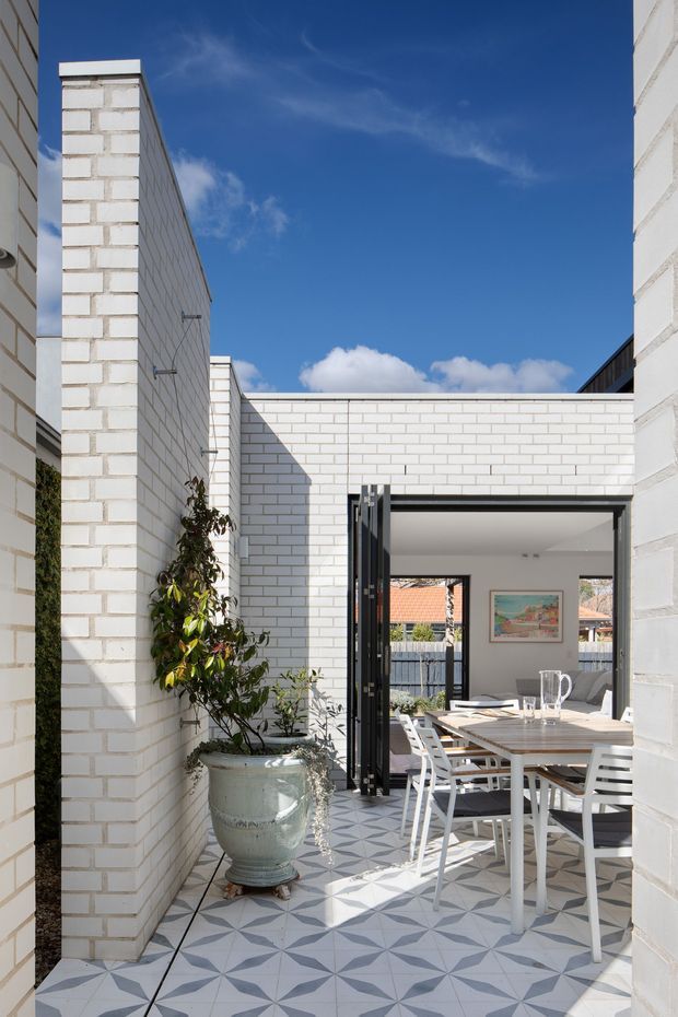 A bright open-air courtyard or terrace enclosed by white brick walls, with a patterned geometric tiled floor, large ceramic planter, and a white outdoor dining table and chairs. Black-framed folding or stacking doors open into a light-filled dining area, creating a strong indoor-outdoor connection. Blue sky and neighbouring rooftops are visible above the walls.