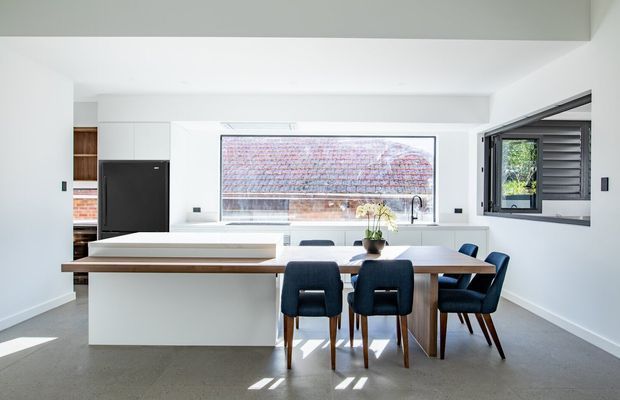 Bright contemporary open-plan kitchen and dining area with a large white island, dark integrated cabinetry, timber dining table, upholstered dining chairs, and expansive windows. The room has a clean white palette, polished concrete flooring, and strong natural light.