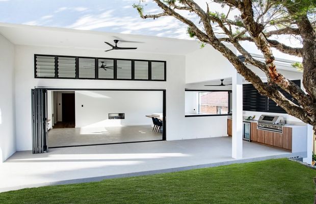 Bright contemporary open-plan indoor-outdoor living space viewed from a landscaped lawn. The white pavilion has extensive black-framed louvred and glazed openings, a ceiling fan, polished concrete flooring, and a wide folding or stacking door opening into an interior living area. A built-in outdoor kitchen with timber cabinetry, dark benchtop, barbecue and sink is visible on the right, while a mature tree extends across the foreground.