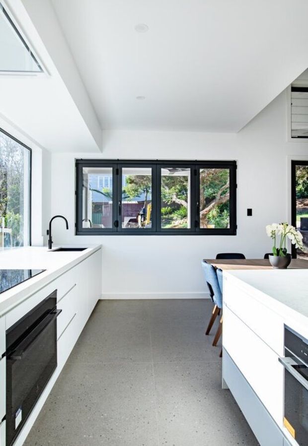 Bright contemporary kitchen with white full-height cabinetry and benchtops, black-framed windows and doors, integrated dark appliances, a central dining area, and a polished grey concrete or terrazzo-style floor. Natural light enters through large glazing and a skylight.