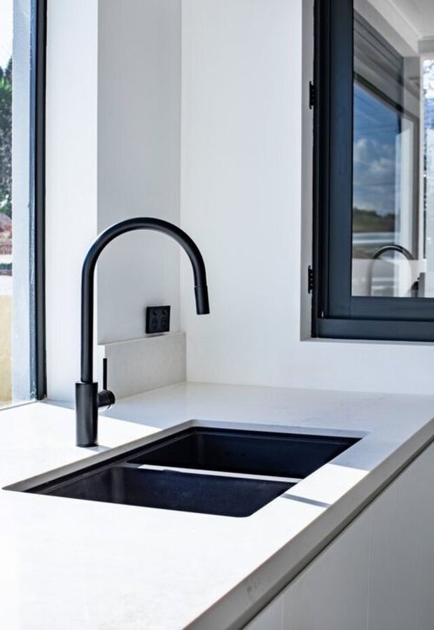 Close-up of a contemporary kitchen sink area with a matte black gooseneck mixer tap, deep black undermount double-bowl sink, white stone-look benchtop, white walls and cabinetry, and dark-framed windows providing abundant natural light.