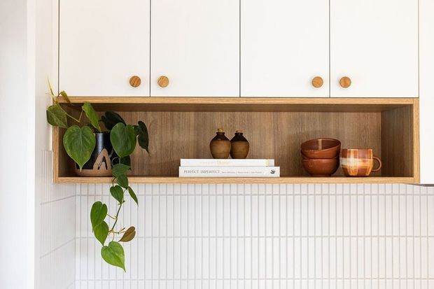 A built-in kitchen or dining-area display niche with white overhead cabinetry, timber-lined open shelving, and a white vertically grooved tile backsplash. The shelf contains ceramic vessels, books, a mug, and a trailing indoor plant, creating a warm natural accent against the light neutral fit-out.