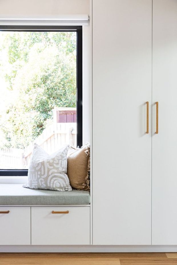 Bright, compact built-in window seat with upholstered cushioning and decorative pillows, integrated beneath a large black-framed window. Adjacent floor-to-ceiling white cabinetry features simple vertical timber handles, with matching drawers below the seat. Warm timber flooring and a leafy outdoor view complete the clean, contemporary interior.