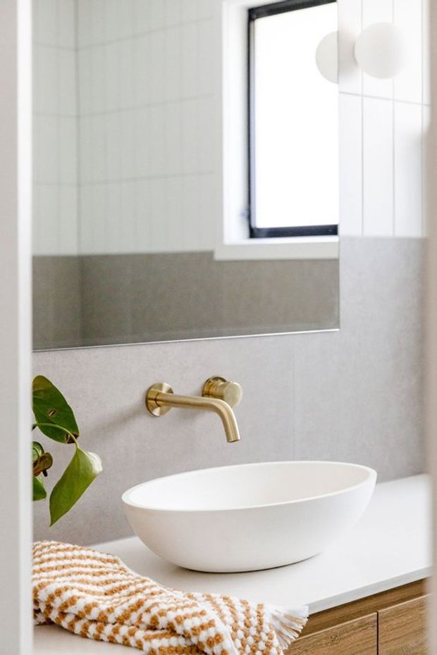 Bright, close-up view of a contemporary bathroom vanity. A white vessel basin sits on a white countertop above warm timber cabinetry, paired with a wall-mounted brushed brass tap. Large mirror, pale grey wall tiles, a small window, and a folded textured towel with a leafy plant create a clean, warm finish.