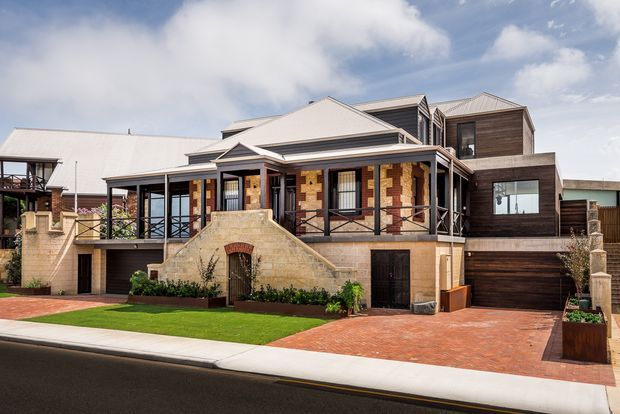 Street-facing exterior view of a substantial multi-level home with a light pitched metal roof, dark timber cladding, sandstone masonry, and prominent covered verandas. The property features raised terraces with dark balustrades, broad steps, a double garage, brick-paved driveway, landscaped front lawn, and contemporary additions integrated with a more traditional residential form.