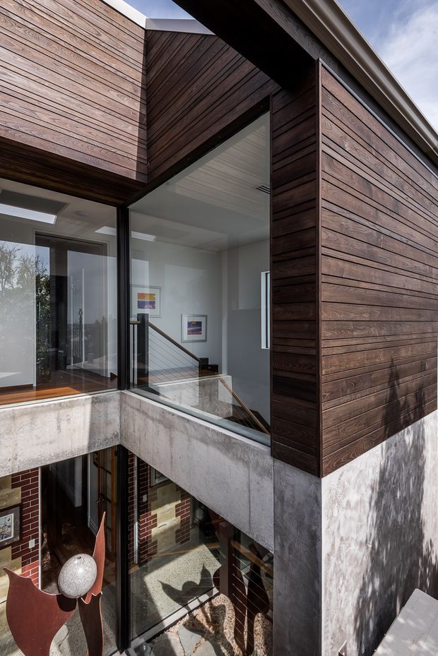 A close architectural view of a multi-level contemporary building focused on a projecting upper volume clad in dark horizontal timber boards. Large glazed openings reveal a bright interior stair landing with framed artwork and a metal handrail. Raw concrete balcony and structural edges contrast with timber cladding, white interior surfaces, and visible brickwork on the lower level. The composition highlights the facade junction, glazing, cantilevered form, and vertical courtyard-like space.