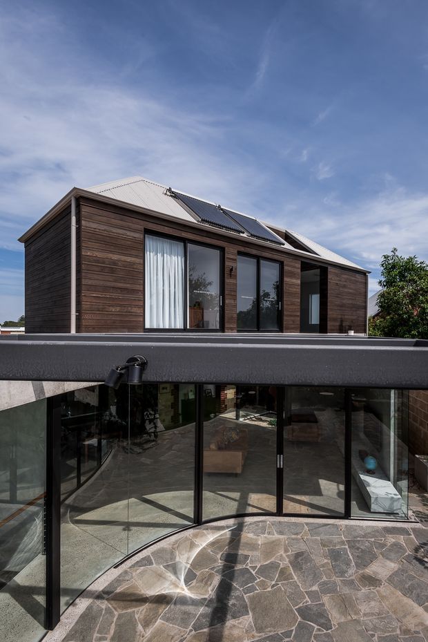 Two-storey residential extension viewed from an elevated courtyard or terrace. The upper level has dark horizontal timber cladding, large black-framed windows and a light corrugated metal pitched roof with rooftop solar panels. The lower level is enclosed by extensive black-framed glazing, opening onto an irregular natural-stone paved courtyard. The image highlights the building facade, glazing and relationship between the upper volume and lower outdoor living area.