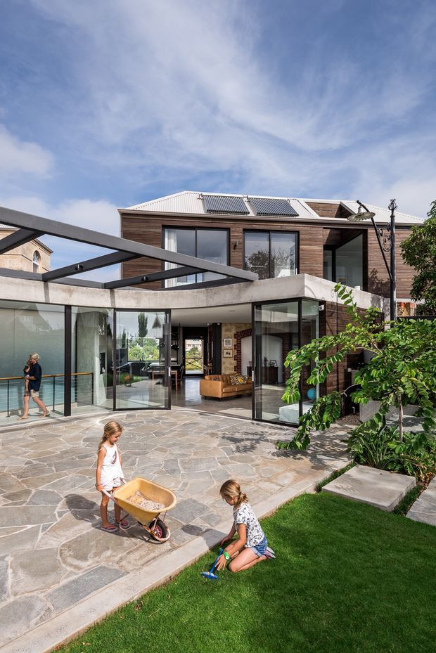 Exterior view of a contemporary multi-level home with dark horizontal timber cladding, extensive floor-to-ceiling glazing, a concrete terrace canopy, and a large paved courtyard. Solar panels are visible on the corrugated metal roof. The open glazing reveals a modern living area, while landscaped planting and lawn surround the patio.