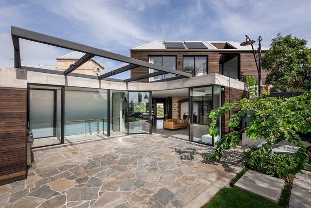Contemporary multi-level residential home viewed from a paved courtyard. The house features extensive floor-to-ceiling glazing, dark timber cladding, exposed angular steel or concrete canopy beams, a flat roof, and a partially enclosed outdoor living area. Solar panels are visible on the upper roof, while established planting borders the courtyard.