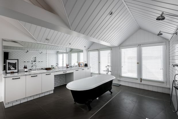 A bright, spacious bathroom with a dramatic white vaulted, panelled ceiling and exposed beams. The room features a freestanding black-and-white bathtub, white cabinetry and vanity joinery, white subway-tile walls, dark large-format floor tiles, extensive louvered windows, wall-mounted shower fixtures, and a minimalist monochrome palette.