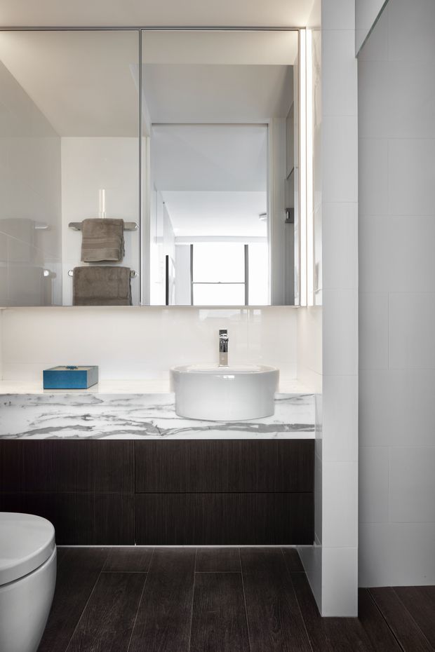 A bright, contemporary bathroom with a wall-mounted dark timber vanity, white vessel basin, chrome tapware, and a white marble-look benchtop. A large mirror spans the vanity, reflecting a towel rail and bright adjacent space. Dark wood-look floor tiles contrast with the white tiled walls and sanitaryware.