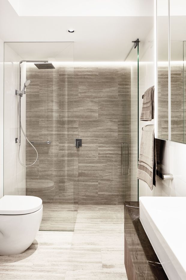 Bright contemporary bathroom featuring a frameless glass walk-in shower, large-format grey-brown stone-look wall and floor tiles, white toilet and bathtub, mirrored cabinetry, integrated lighting, and chrome and dark metal fixtures.