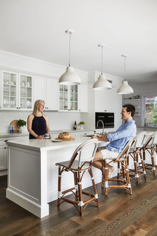 Bright, refined kitchen interior with a large white island and integrated seating, white glass-front cabinetry, pale countertops, dark timber flooring, and three oversized white pendant lights. Two people are present at the island, with woven rattan bar stools providing a warm natural contrast. The room has traditional moulded ceiling trim and a calm, airy coastal-Hamptons character.