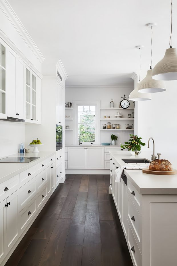 Bright, narrow galley-style kitchen with extensive white Shaker cabinetry, dark timber flooring, white benchtops, open shelving, integrated appliances, a central island with sink, and three oversized ribbed pendant lights. Traditional detailing includes ornate cornices, framed cabinetry, cup pulls, and a large wall clock, creating a refined classic look.
