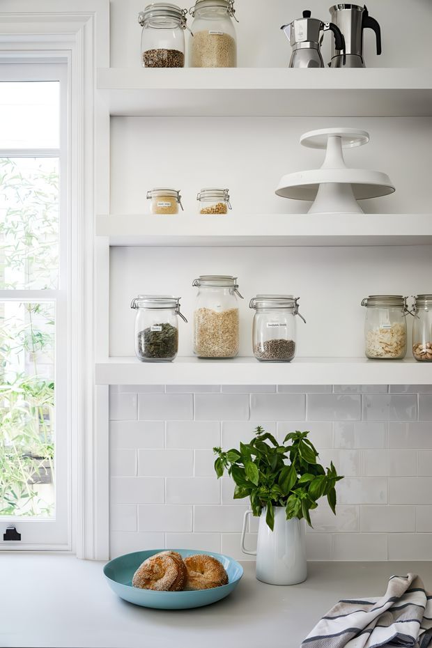 Bright, neatly styled kitchen pantry area with open white shelving displaying labelled glass storage jars, coffee makers, and a white cake stand. A white subway-tile splashback sits above a pale countertop, with a leafy plant in a white jug, a bowl of bread, and a striped tea towel. A tall white-framed window brings in abundant natural light.