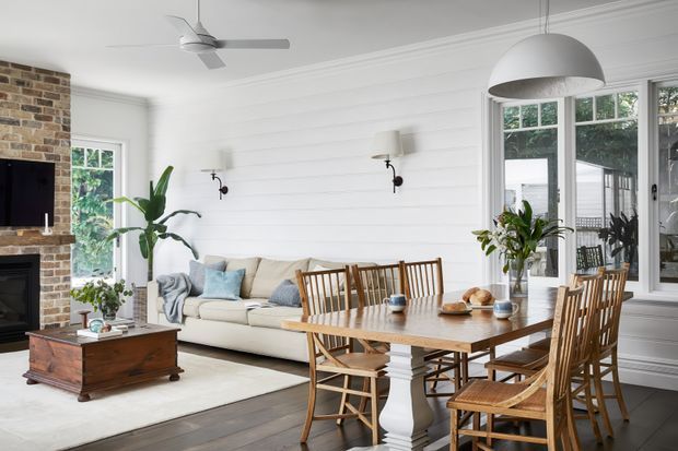 Bright open-plan living and dining room with a relaxed coastal-traditional character. The room features white horizontal wall panelling, white-painted trim, a red-brown exposed brick fireplace wall, dark timber flooring, a cream sofa, and a substantial timber dining table with spindle-back chairs. Large multi-pane windows bring in abundant natural light, while a white ceiling fan and oversized dome pendant provide practical and decorative lighting. Indoor plants, soft blue cushions, and neutral furnishings create a fresh, inviting atmosphere.