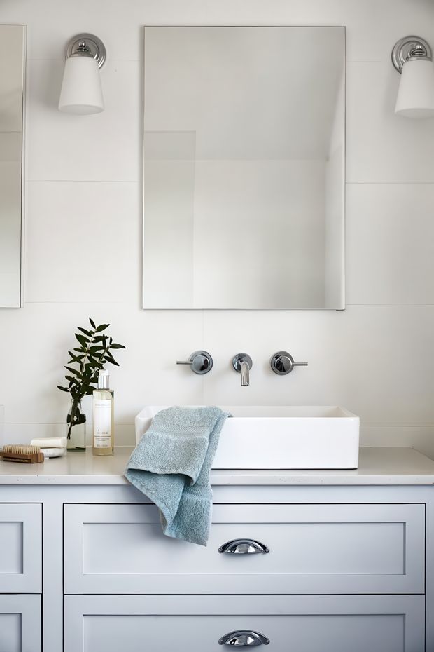 A bright, refined bathroom vanity scene with a large frameless rectangular mirror, chrome wall-mounted faucet, white vessel basin, integrated pale countertop, and light grey-blue paneled vanity cabinetry with polished chrome pulls. Matching chrome-and-white wall sconces flank the mirror. A folded pale blue towel, soap dispenser, and small leafy plant add restrained styling.