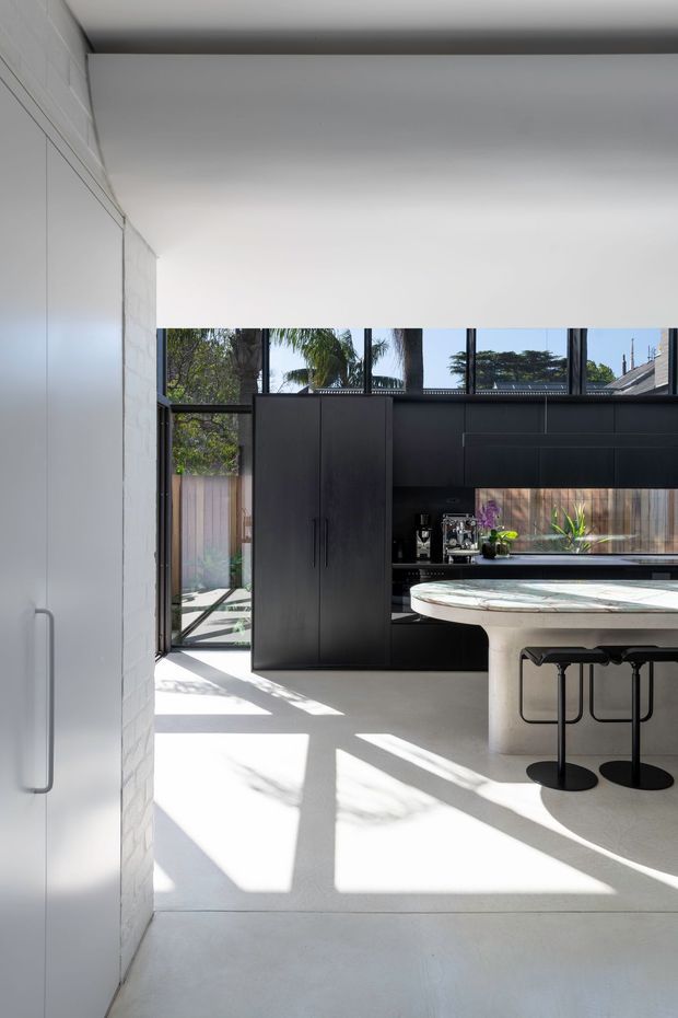 A bright, contemporary kitchen with a minimalist black built-in cabinetry wall, tall integrated storage, dark-framed clerestory glazing, and a large curved white stone island with bar stools. Sunlight casts strong geometric shadows across the pale floor, while a glazed door opens toward a planted outdoor area.