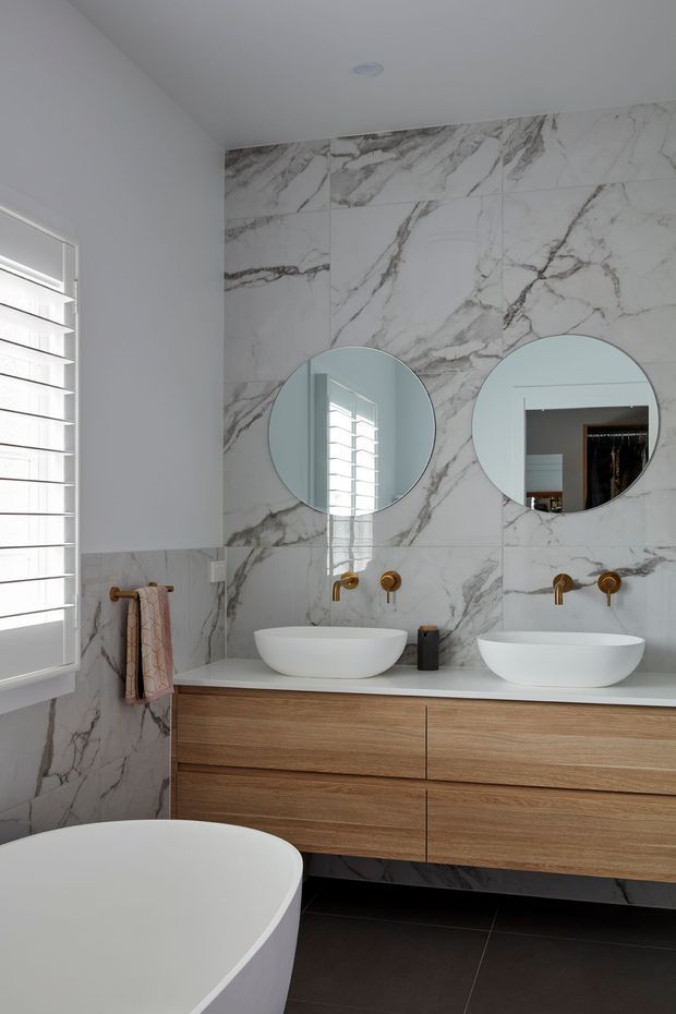 Bright contemporary bathroom featuring a double floating timber vanity with white vessel basins, wall-mounted brass tapware, two round mirrors, and a full-height white marble-look veined wall finish. A freestanding white bath is partially visible in the foreground, with dark floor tiles and plantation shutters adding contrast.