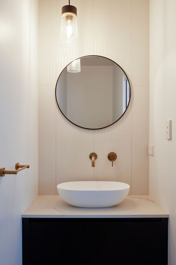 A compact, refined bathroom vanity photographed straight on. The room features a white vessel basin on a pale marble-look benchtop, dark integrated vanity cabinetry, brushed brass wall-mounted tapware, a large round mirror with a dark thin frame, and a pendant light with a clear glass shade. Vertical textured wall panelling provides a subtle feature backdrop.