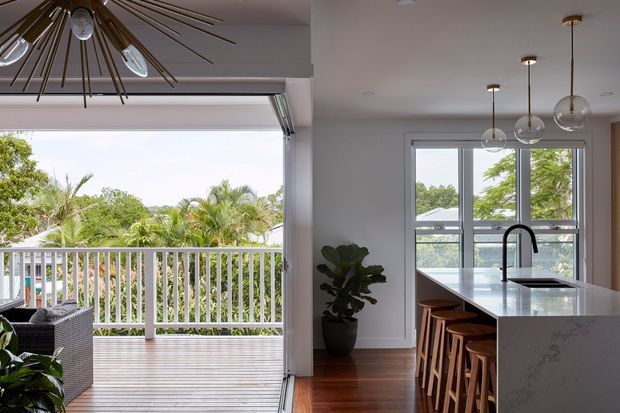 Bright open-plan kitchen and living area with a large sliding glass opening to a timber deck and leafy outdoor setting. The kitchen features a long white stone island with seating for several people, dark cabinetry, a black gooseneck tap, timber flooring, and contemporary pendant lighting. A woven outdoor seating area is visible beside the deck.