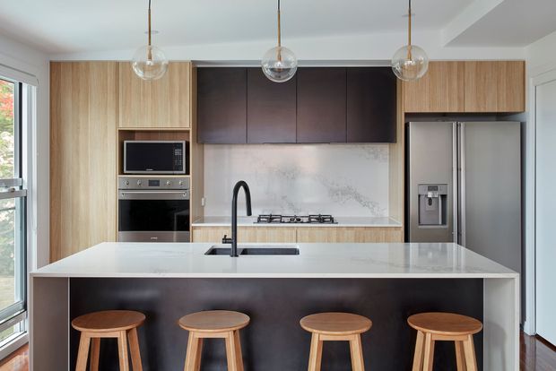 Bright contemporary kitchen featuring a large white island with integrated sink, dark contrasting island cabinetry, four timber stools, pale timber cabinetry, dark upper cupboards, stainless-steel appliances, a pale stone-look splashback, and three glass pendant lights. Large glazing at the side brings in natural light.