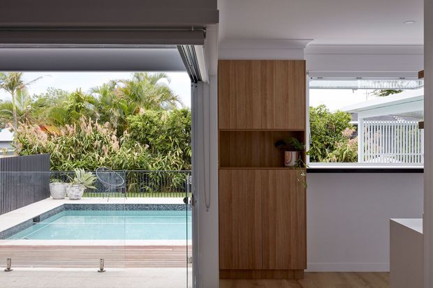 Bright contemporary interior opening directly onto a swimming pool and lush tropical garden. Large sliding glass doors create a seamless indoor-outdoor connection, while a full-height warm timber storage partition with open shelving divides the space. White walls, a pale ceiling, timber flooring, and a partial light-coloured kitchen or cabinetry element are visible.