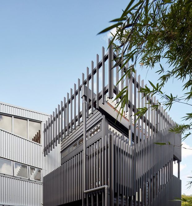 Close-up exterior view of a contemporary multi-storey building with corrugated metal cladding, extensive vertical metal fins, and layered screened balconies or circulation spaces. The facade has a strong geometric, industrial character, with adjacent glazing and bamboo-like planting partially framing the view.