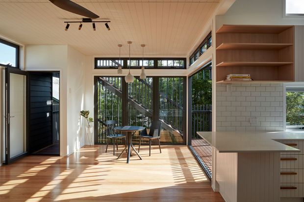 A bright, open-plan kitchen and dining area with extensive black-framed glazing, a timber-lined ceiling, pale timber flooring, and a compact dining table beside the windows. The kitchen features white vertical-groove cabinetry, a light benchtop, white subway-tile splashback, and open timber shelving. Strong sunlight casts linear shadows across the floor, while an exterior stair and greenery are visible through the glazing.