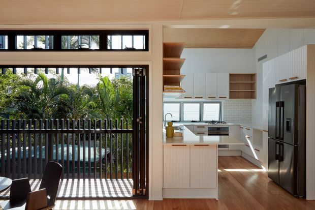 Bright contemporary kitchen opening onto a timber deck or balcony through large dark-framed sliding doors. The kitchen features white flat-panel cabinetry, open timber shelving, a pale tiled splashback, timber flooring, stainless-steel appliances, and a central island. Tropical planting is visible outside, with strong linear sunlight and shadows across the deck and floor.
