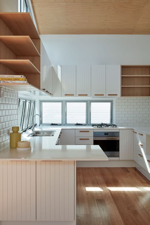 Bright, compact contemporary kitchen with white vertical-panel cabinetry, open timber shelving, white subway-tile splashback, pale stone benchtops, stainless-steel sink and integrated oven. Warm timber flooring and a timber-lined ceiling add natural contrast, while large windows provide strong daylight. The layout includes a substantial island and a wall of storage cabinetry.