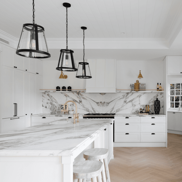 Bright, refined white kitchen with extensive marble-look surfaces, a large waterfall island, white shaker-style cabinetry, brass tapware and hardware, and three black-framed pendant lights. The room includes a marble backsplash, rangehood, integrated appliances, and a warm herringbone timber floor.
