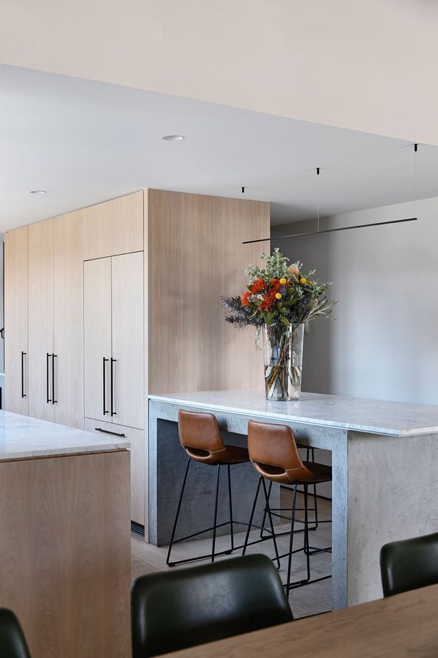 Contemporary kitchen interior featuring full-height light timber cabinetry, a large pale stone island and peninsula with waterfall edge, dark integrated appliances or panels, and a simple white ceiling with recessed and linear lighting. Two brown leather bar stools sit at the island, with dark dining chairs and a timber dining table in the foreground. A colourful floral arrangement provides a small accent against the restrained neutral palette.