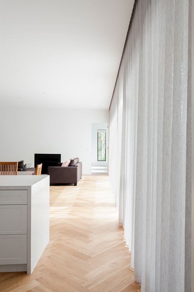 Bright, minimalist open-plan interior viewed along a wall of full-height sheer curtains. Pale herringbone timber flooring leads toward a compact living area with a dark fireplace, brown sofa, and timber dining chairs. A white kitchen island with integrated drawers occupies the foreground, while a narrow glazed doorway provides additional natural light at the rear.