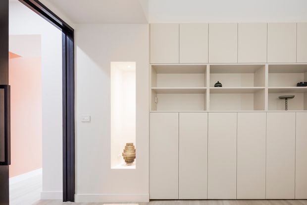 Minimal contemporary interior with a full-height built-in storage and display wall in a warm off-white finish. The joinery combines tall closed cupboards with open shelving and recessed niches, paired with dark-framed glazing or a doorway at left. A small decorative object sits in the illuminated niche, and a glimpse of an adjacent room adds a soft blush accent.
