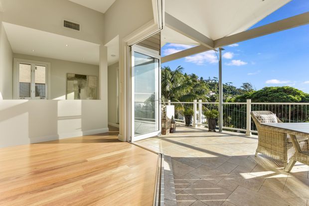 Bright open-plan interior opening directly onto a covered outdoor terrace through wide glazed folding or sliding doors. The room has pale walls, light timber flooring, a simple white kitchen or service counter at left, and a tiled terrace furnished with wicker dining chairs and a table. White railings, slim posts, tropical vegetation, and blue sky are visible beyond the terrace, creating a strong indoor-outdoor connection.
