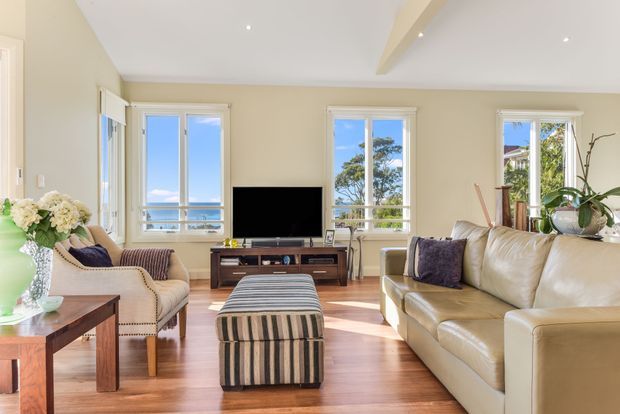 A bright, comfortably furnished living room with cream walls, polished timber flooring, large white-framed windows, and a pale leather sofa. Two upholstered armchairs flank a timber coffee table, while a striped ottoman sits centrally facing a television and low timber media console. Indoor plants and floral arrangements add natural detail, and the room overlooks a sunny exterior through the windows.