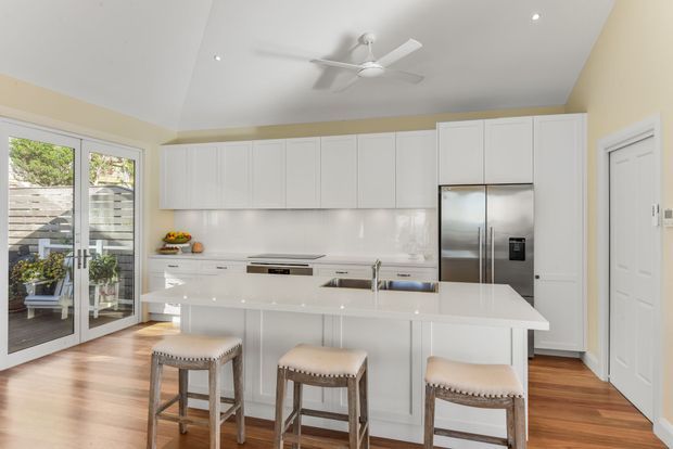Bright, spacious contemporary kitchen with extensive white Shaker-style cabinetry, a large white island with integrated sink and seating for three, stainless-steel French-door refrigerator, pale reflective splashback, and warm timber flooring. Full-height glazed French doors open to an outdoor area, while recessed downlights and a white ceiling fan provide lighting and ventilation.