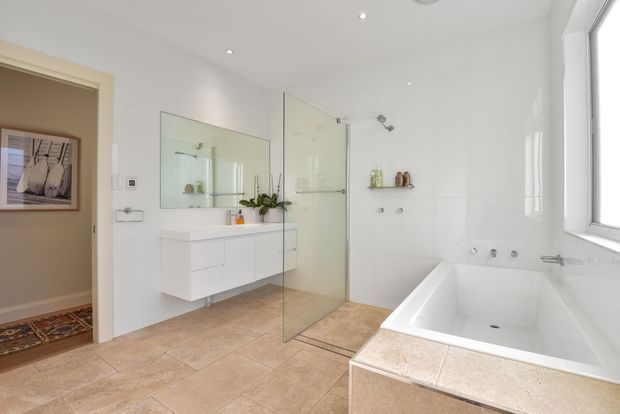 Bright contemporary bathroom with a large white freestanding-style bathtub set into a tiled surround, frameless glass shower enclosure, floating white vanity with drawers, wall-mounted mirror, and pale stone-look floor and wall tiles. A doorway opens to an adjacent carpeted room, while a large window provides natural light.