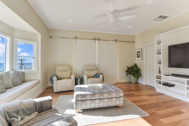 Bright, spacious living room with warm timber flooring, pale neutral walls, a built-in window seat, two upholstered armchairs, a patterned ottoman, area rug, ceiling fan, and white built-in entertainment shelving. Large windows bring in abundant daylight and offer a water view. The room has a relaxed, coastal-influenced contemporary feel.
