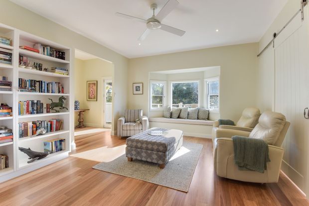 Bright, traditional-style living room with warm timber flooring, pale sage walls, built-in white bookcases, a window seat in a bay-style glazed alcove, upholstered armchairs, a patterned ottoman, and a ceiling fan. The room has abundant natural light and a comfortable, inviting atmosphere.