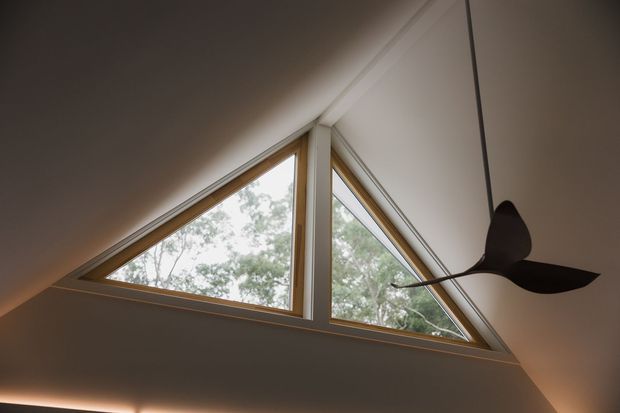 Interior architectural detail showing a high sloped ceiling with a large triangular clerestory window assembly. Warm timber-toned window frames contrast with pale plastered surfaces, and a sculptural dark pendant light hangs in the foreground. Trees are visible outside through the glazing.
