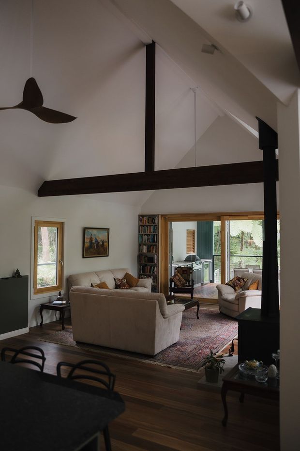 A warm, high-ceilinged living room with exposed dark timber beams and a ceiling fan. The room features white walls, dark timber flooring, a large patterned rug, upholstered sofas and armchairs, built-in bookshelves, and timber-framed windows and sliding doors opening to a bright outdoor deck. A dining table is partially visible in the foreground.