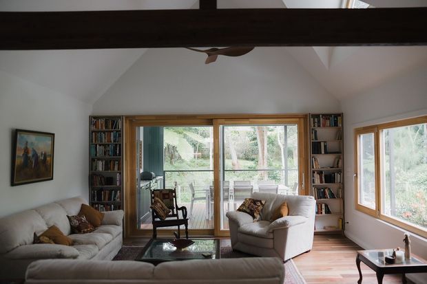 A bright, cozy living room with a vaulted white ceiling, exposed dark timber beam, large timber-framed windows and glazed doors, built-in bookshelves, upholstered sofas and armchairs, and a view toward a leafy outdoor setting. Timber flooring, a glass coffee table, artwork, and layered cushions add warmth and character.