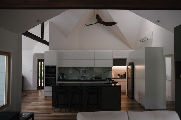 Contemporary open-plan kitchen viewed from a living area, featuring a dark kitchen island with bar seating, white full-height cabinetry, integrated appliances, a dark stone or glass backsplash, timber flooring, and a dramatic vaulted ceiling with a ceiling fan. Adjacent living and entry areas are visible.