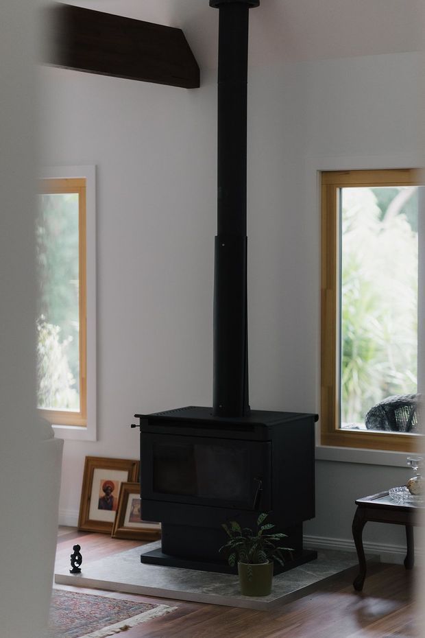 Interior view of a cozy room centred on a freestanding black wood-burning stove with a tall flue pipe. White walls, timber-framed windows, exposed dark timber ceiling beam, hardwood flooring, framed artwork, a patterned rug and traditional dark furniture create a warm, rustic setting.