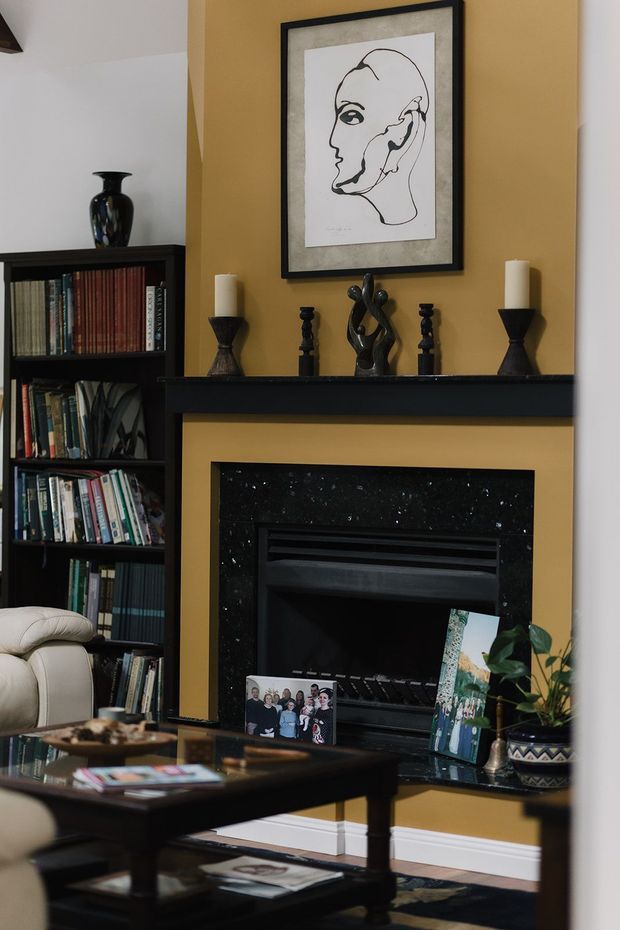 A traditional living room centered on a black fireplace with a mustard-gold accent wall and substantial black mantel. The mantel is styled with candlesticks and a sculptural ornament, while a framed line-art portrait hangs above. A tall dark bookcase filled with books stands to the left, and a dark wood coffee table, upholstered seating, framed family photographs, and a potted plant complete the warm, classic interior.
