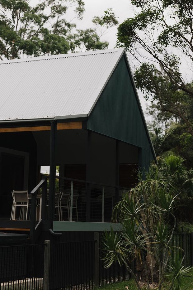 Exterior view of a compact contemporary house set among mature vegetation. The building features a steep gable roof clad in light corrugated metal, dark green vertical ribbed cladding, deep covered verandah areas, dark structural framing, timber soffit accents, and a raised outdoor deck or balcony with metal balustrading. Lush planting surrounds the fenced property.
